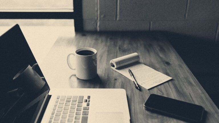 Laptop, coffee mug, notepad with pen, and smartphone on a wooden table beside a window in soft morning daylight.
