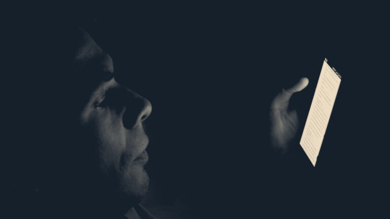 Man's profile lit only by the glow of a smartphone held up in a dark room at night.