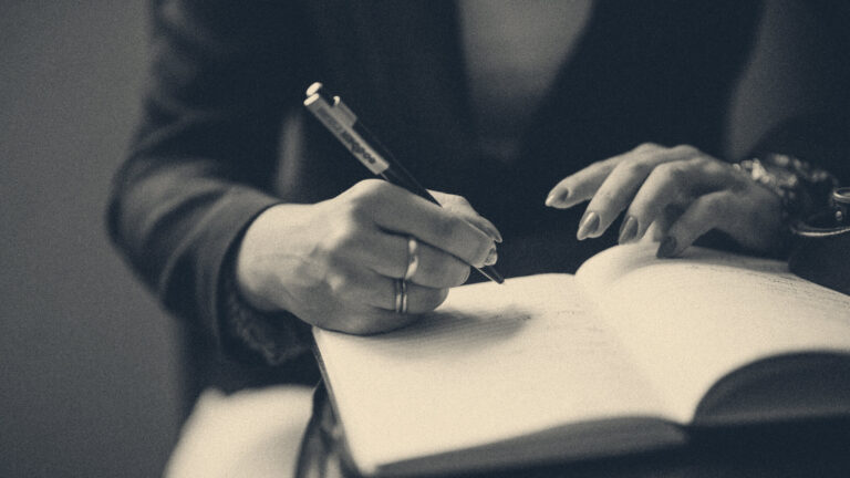 Close-up of a woman's hands writing in an open notebook with a pen, soft warm indoor light
