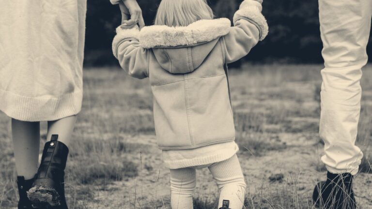 A toddler in a duffel coat walks along a woodland path, held by a parent's hand on each side.