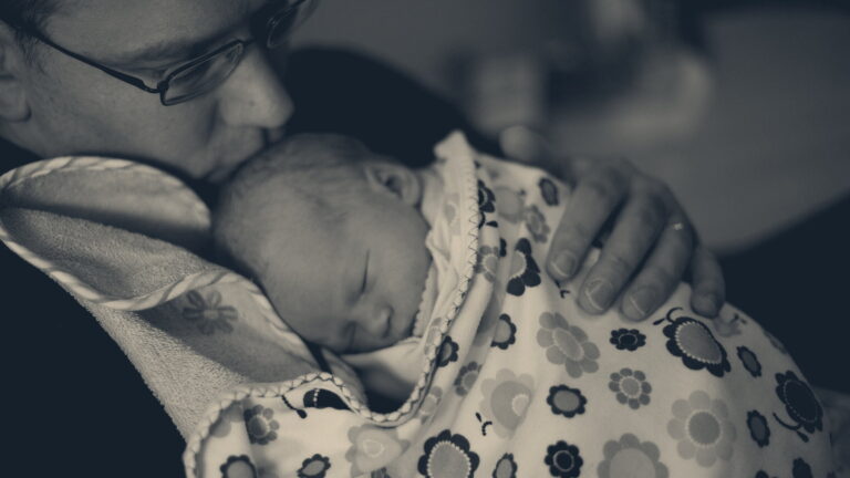 A father in glasses cradles his newborn daughter wrapped in a flower-print blanket, in hospital light