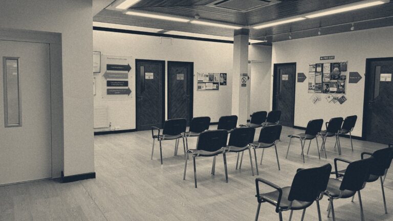 Empty chairs in a British GP surgery waiting room, consulting-room doors behind