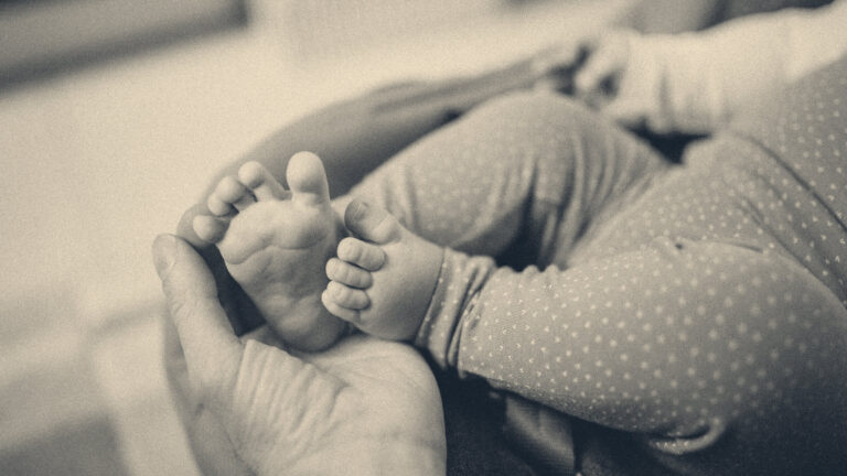 A parent's open hand gently cups the tiny bare feet of a newborn baby resting in a soft patterned bouncer.