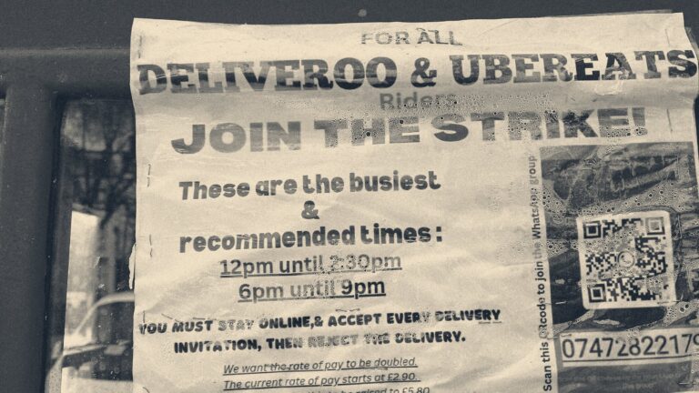 Rain-flecked flyer in a red London phone box urging Deliveroo and UberEats riders to join a strike via a WhatsApp QR code.