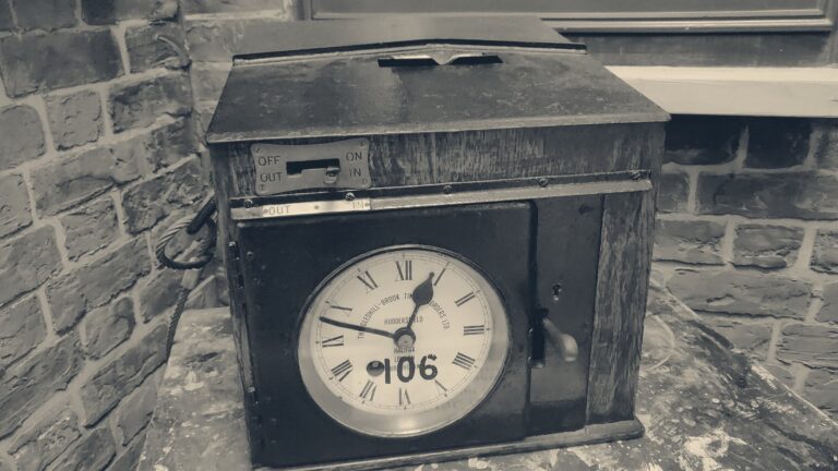 Vintage Gledhill-Brook clocking-in machine with clock face and card slot on a workshop bench against red brick