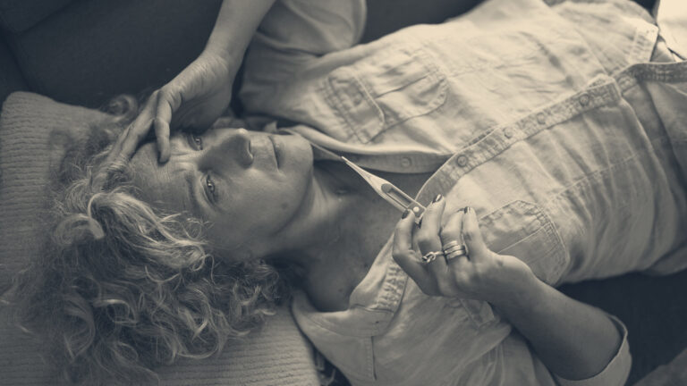 A woman recovering on a sofa checks a digital thermometer, hand resting on her forehead.