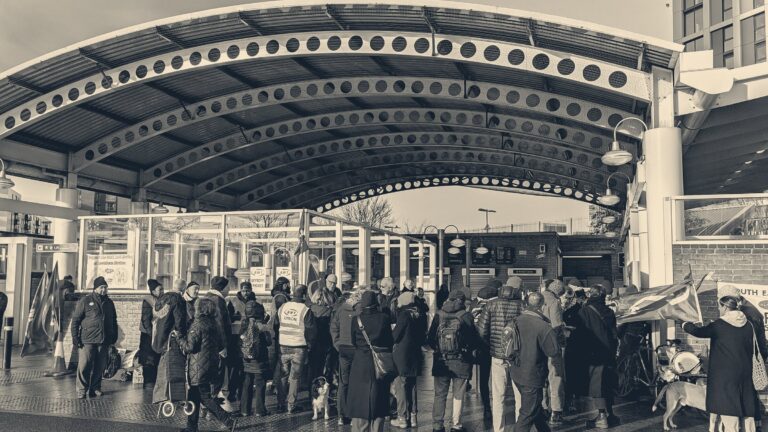 Striking workers in winter coats and an RMT union hi-vis vest gather under the iron canopy outside Lewisham station, London