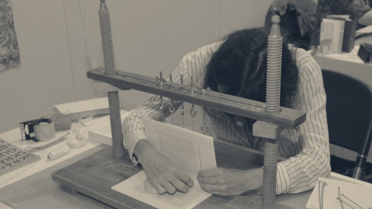Apprentice bent over a wooden sewing frame, stitching a book block by hand at a workbench.