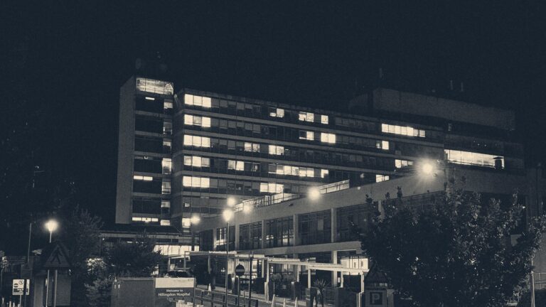 Hillingdon Hospital's brutalist block at night, scattered ward windows lit above the darkened entrance