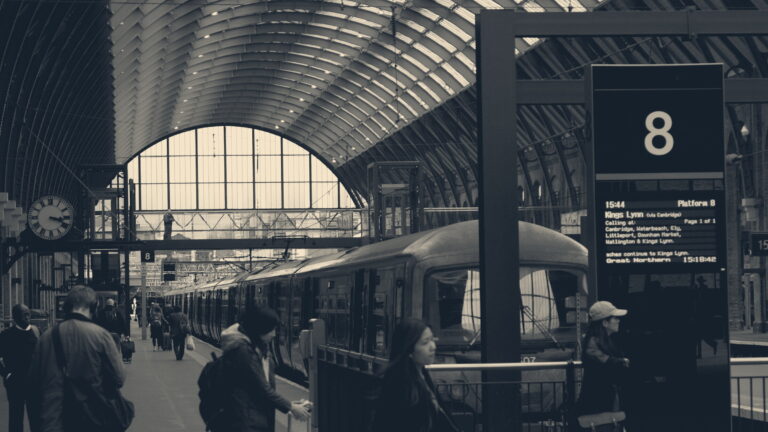 Commuters at London King's Cross under the arched roof, with a platform clock and departures board showing 15:44
