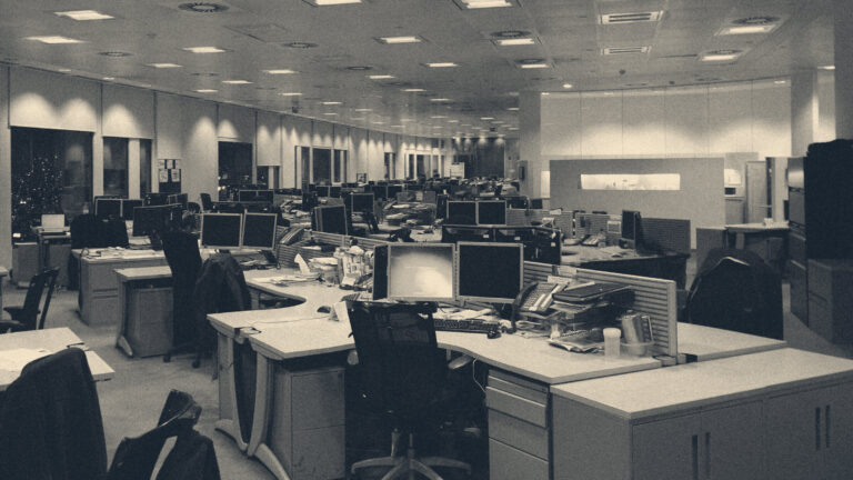 Empty open-plan London office floor at night, with curved desks, paper stacks, and a glowing computer screen.