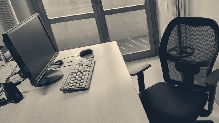 An empty office chair pushed back from a desk with a dark monitor, keyboard and mouse beside tall windows.