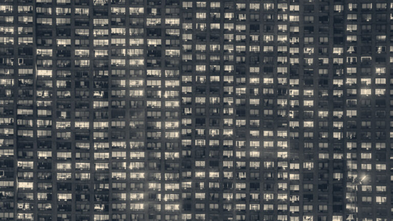 Dense grid of lit and dark windows in a high-rise block of flats at night, a street lamp glowing at lower right