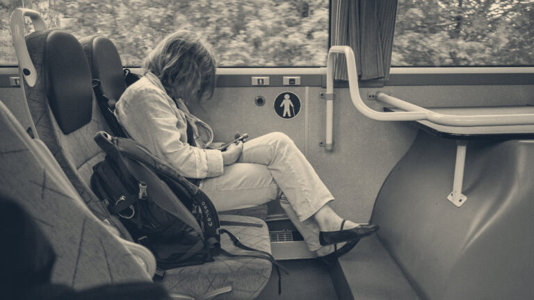 A woman sits alone on a bus seat, absorbed in her smartphone, beside blue seats and a yellow handrail.