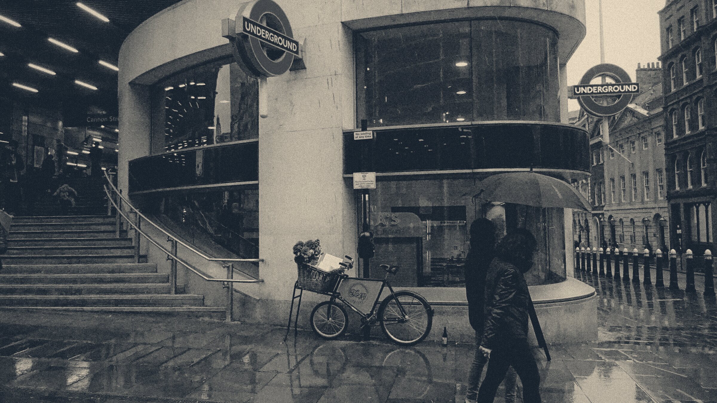 Commuters under umbrellas pass Cannon Street Underground entrance on a rain-slicked City of London street.