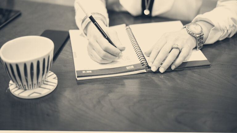 A person's hands write notes in a spiral-bound diary on a wooden desk beside a mug and phone.