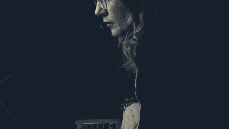 Woman in profile working on a laptop in near-darkness, screen glow lighting her tired face and hand