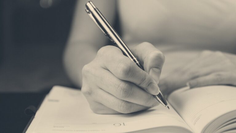 Hand holding a silver pen, writing in a dated desk diary; moody dark background with soft bokeh.