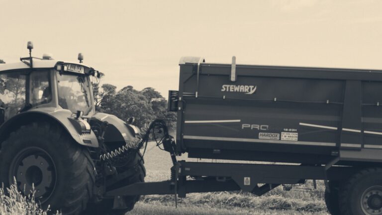 Green tractor towing a blue grain trailer through a golden wheat field at harvest, Moray, Scotland.