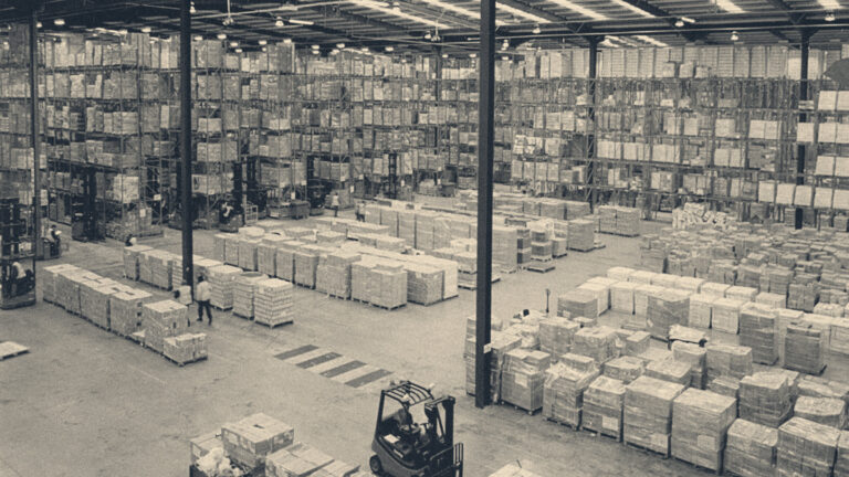 A red forklift truck and hi-vis workers among pallet stacks and towering storage racks in a distribution warehouse.