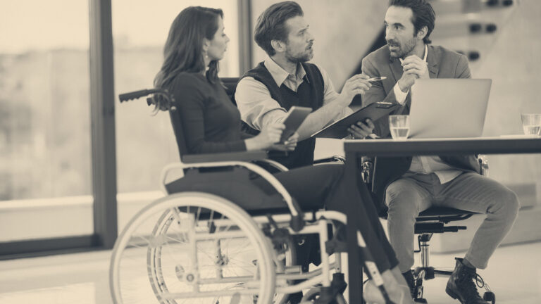 A woman in a wheelchair discussing documents with two colleagues at an office table beside bright windows