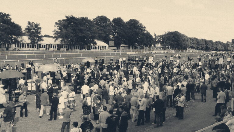 Punters crowd the bookmakers' betting ring at Newmarket's July Course, studying colourful odds boards beside the rails.