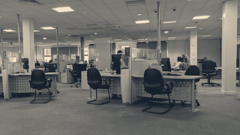 A claimant meets a work coach at pod 12 inside a Cardiff Jobcentre Plus, under fluorescent office lighting.