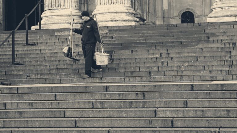 A street cleaner carries a broom and bucket down the wide stone steps of a London landmark.