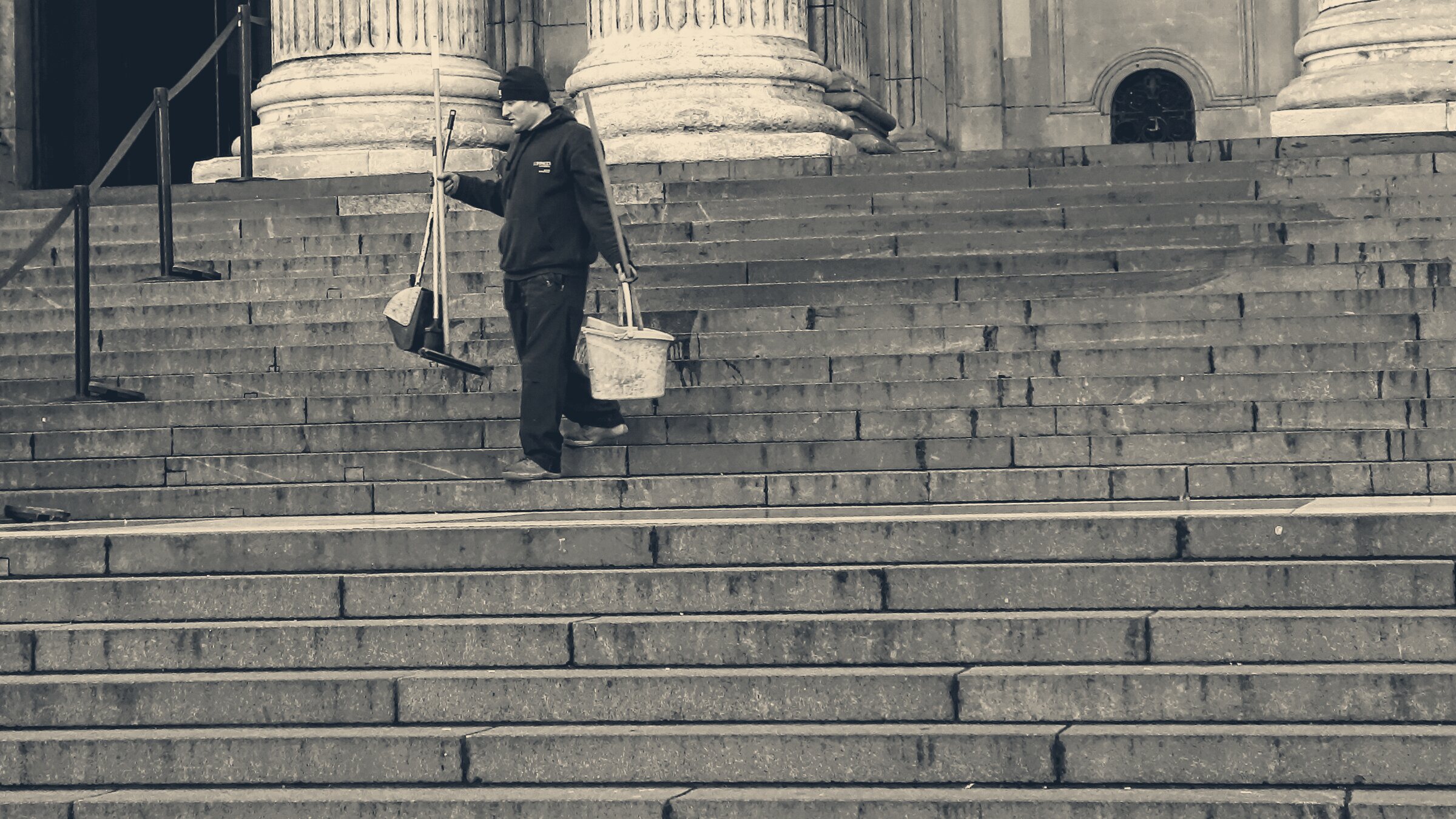 A street cleaner carries a broom and bucket down the wide stone steps of a London landmark.