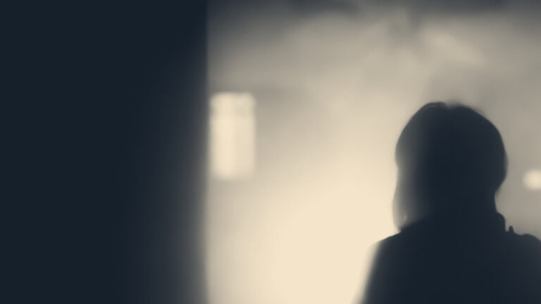 Silhouetted person seen from behind stands in a misty interior beside a dark doorway, facing bright light