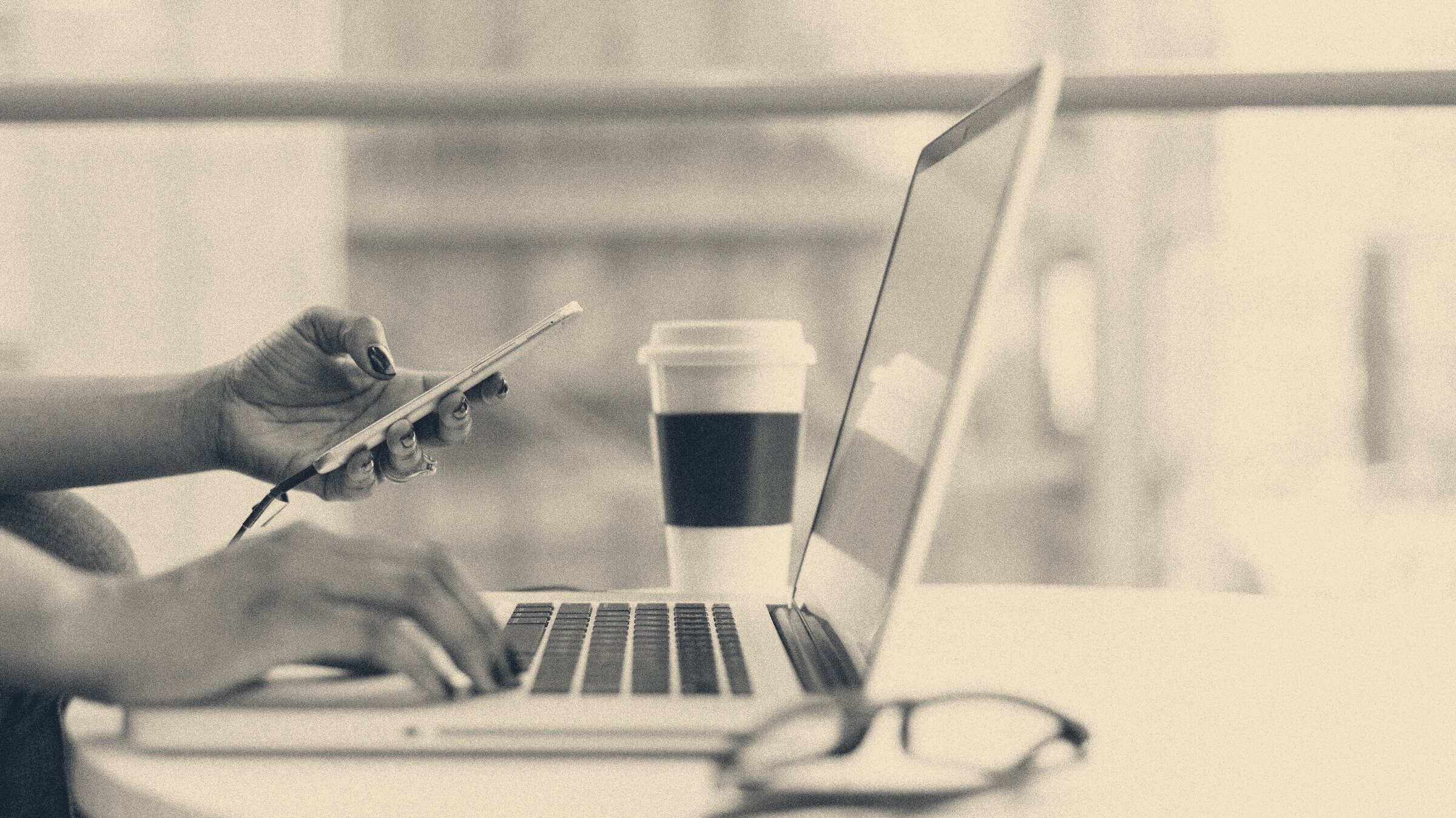Office worker at a desk rests a hand on a laptop keyboard while holding a smartphone beside a takeaway coffee cup.