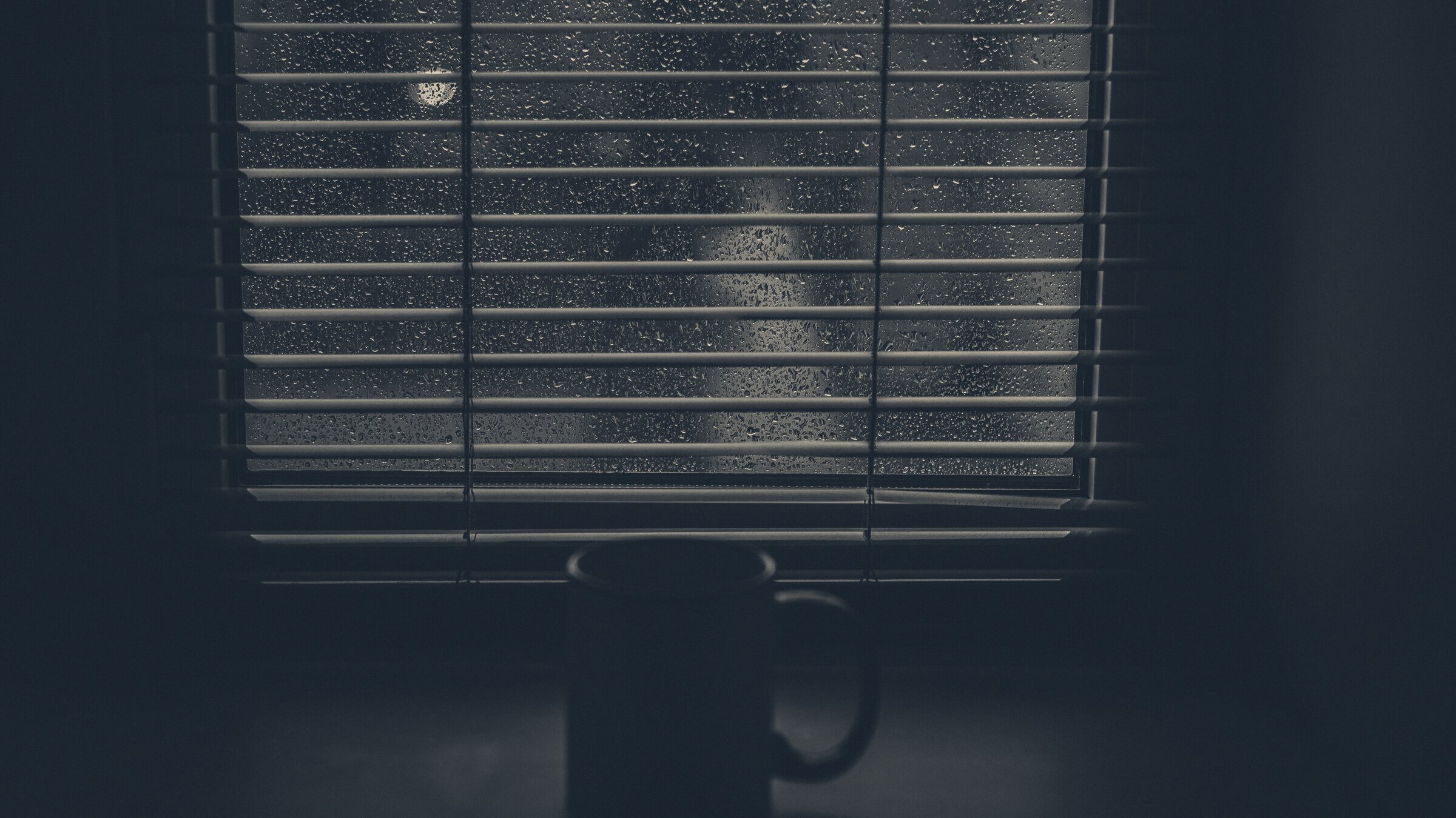A dark mug sits on a windowsill in front of venetian blinds streaked with rain, lit by a distant streetlight.