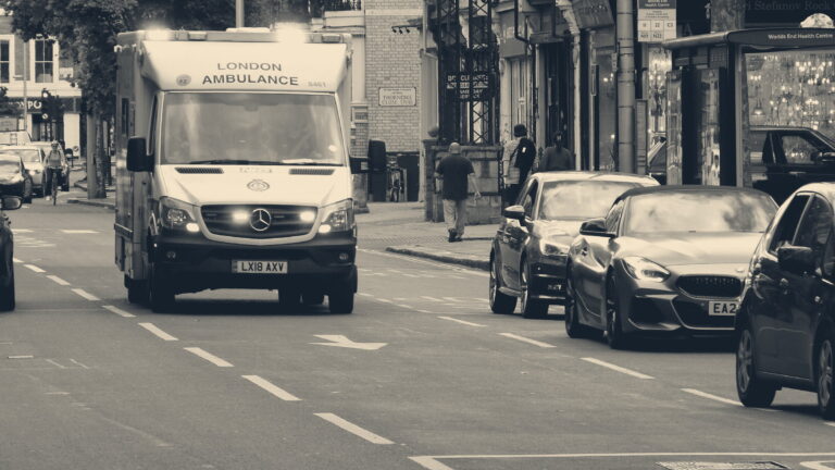 NHS ambulance with blue lights on a Chelsea street, passing the Worlds End Health Centre bus stop