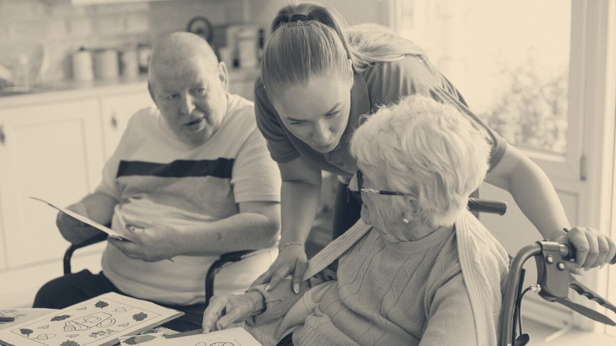 Care worker in blue uniform supports an elderly woman in a wheelchair at a kitchen table during a home visit.