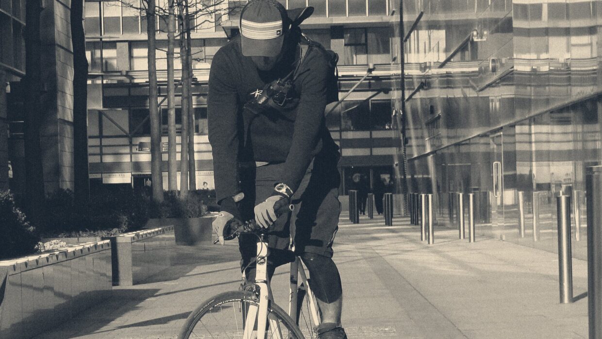Bicycle courier in black cycling gear riding across a London corporate plaza, head down under a cap visor.