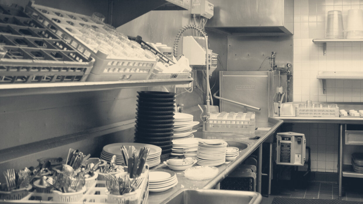 Commercial kitchen dishwashing station with stacked plates, cutlery caddies, and pass-through dishwasher after service