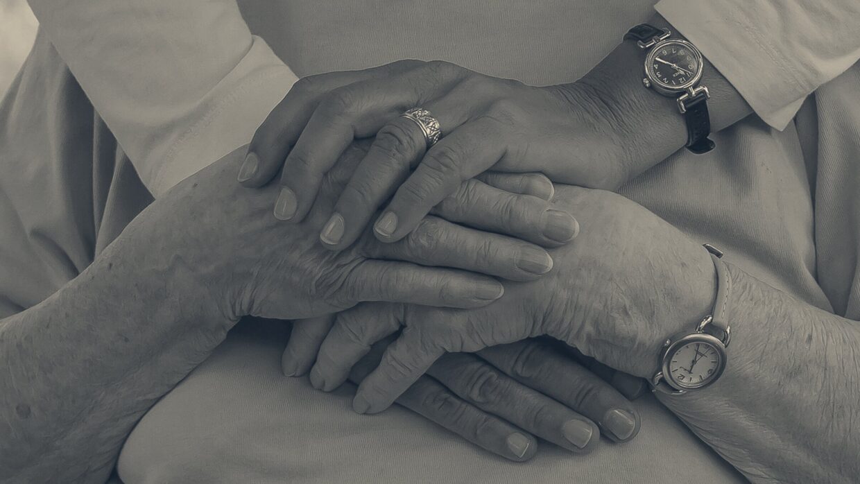 Caregiver embracing an older person from behind with hands clasped together, wristwatches visible on both wrists.