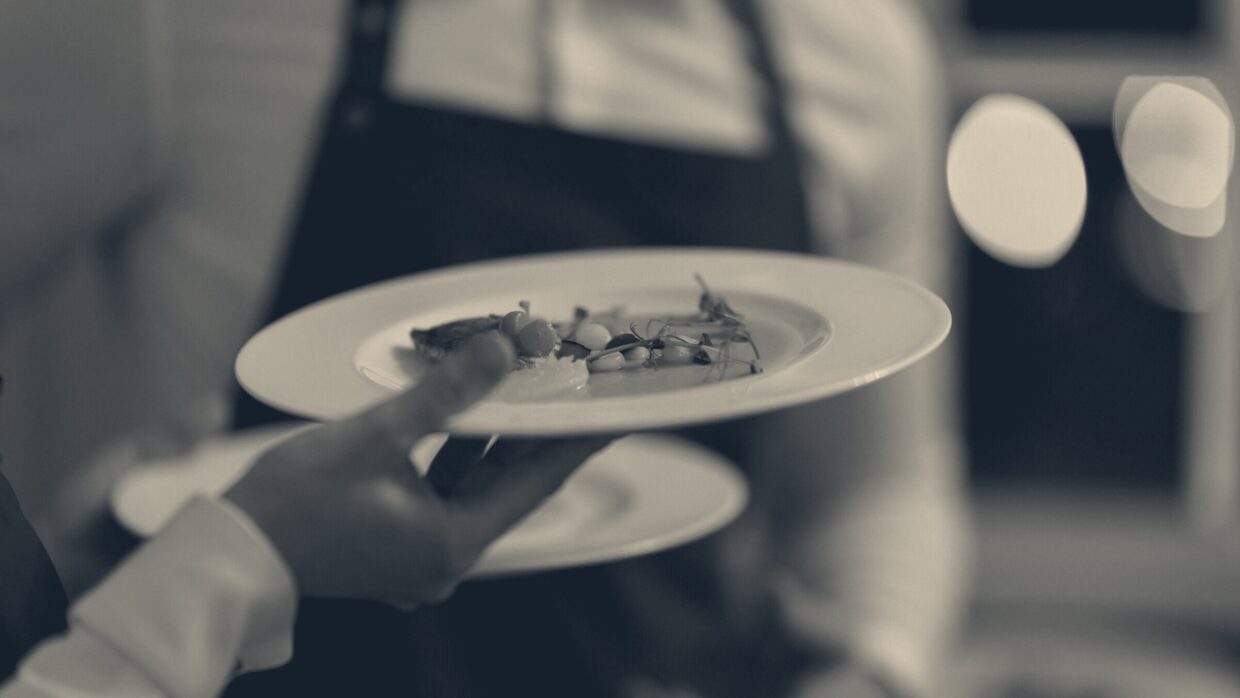 A waiter in a dark apron and white shirt carries dinner plates in a restaurant, with a colleague in the background