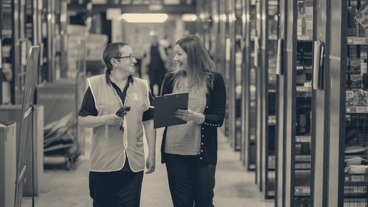 Two warehouse workers talking while walking down an aisle lined with blue storage racking, one holding a clipboard.
