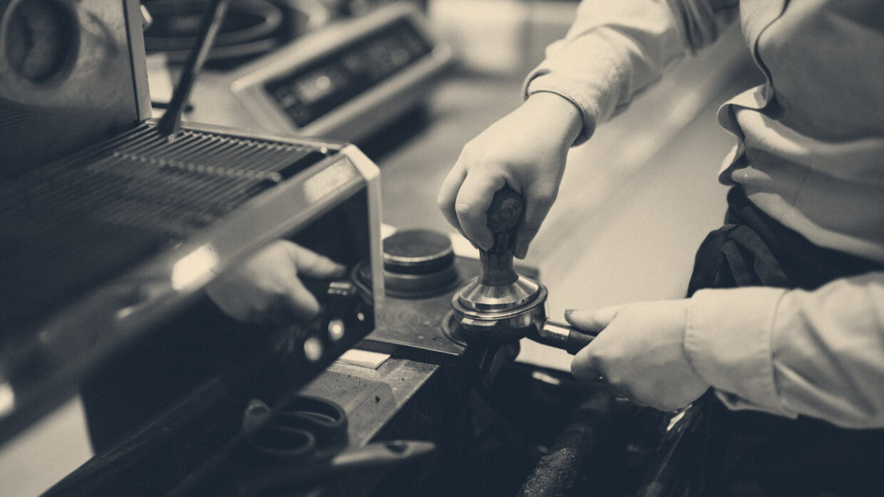 Barista in an apron and blue shirt tamping coffee grounds into a portafilter at a cafe espresso station.